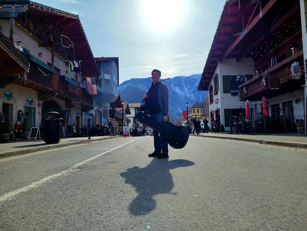 A man with a guitar case stands in the middle of a street lined with alpine-style buildings and mountains behind.