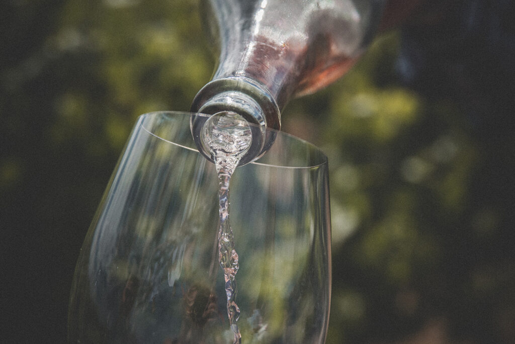 Close-up of a bottle from the Ultimate Evergreen Family Wines Club pouring a clear liquid into a wine glass, with a blurred green background.