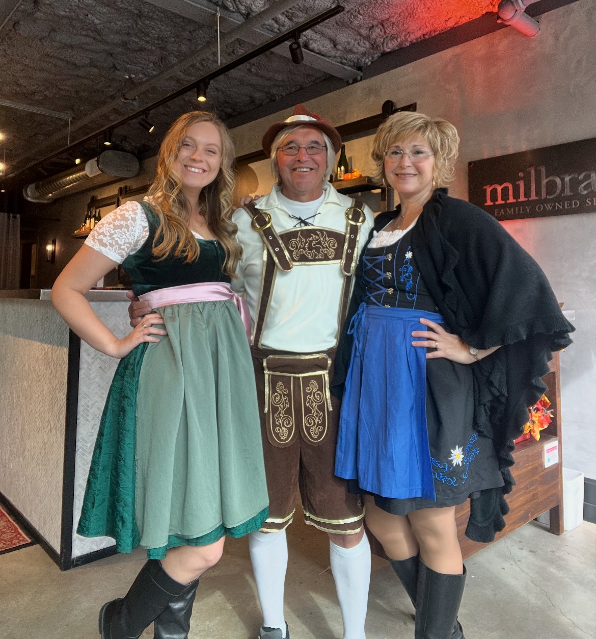Three adults in traditional Bavarian outfits smiling together inside a modern, warmly-lit building.