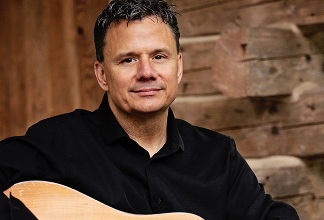 MichaelGood Man with short dark hair in a black shirt holding an acoustic guitar, sitting by a wooden wall.