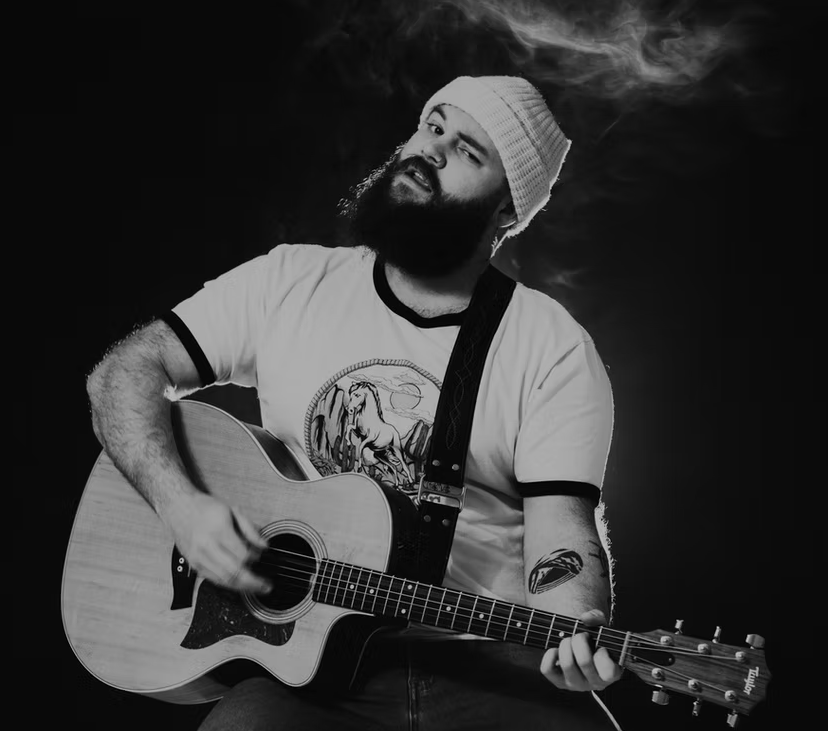 Screenshot 2025-10-21 124530 Bearded man in a beanie playing an acoustic guitar, with smoke swirling in the background, black and white.