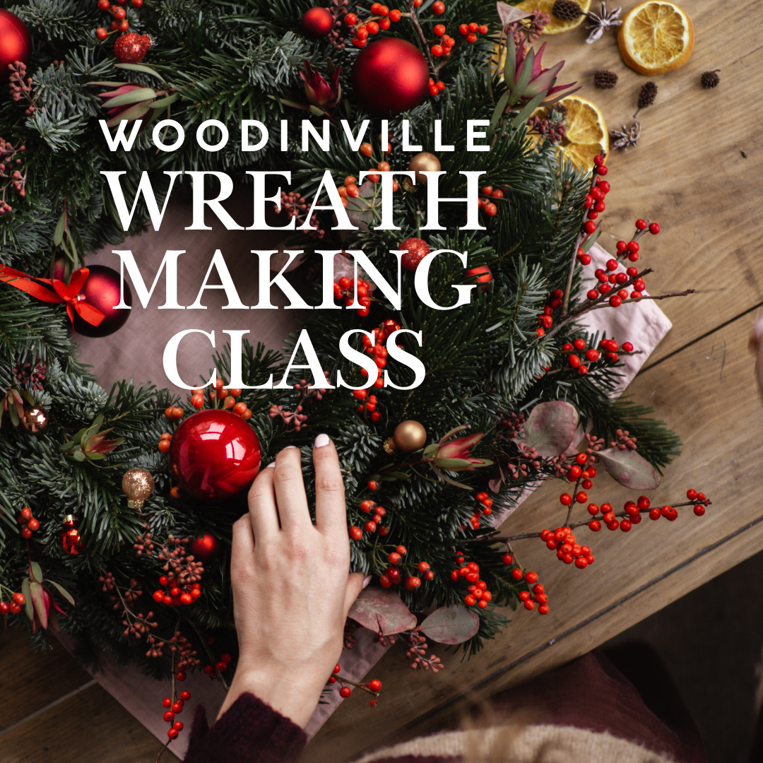 A person arranges a festive wreath with red ornaments and berries; text reads Woodinville Wreath Making Class.