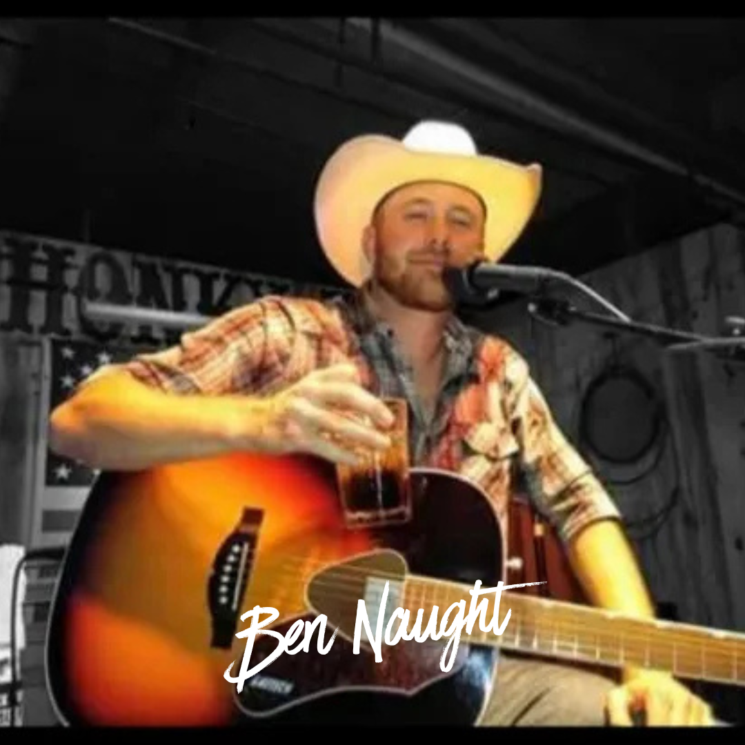 A man in a cowboy hat plays guitar and holds a drink, sitting by a microphone in a rustic setting.