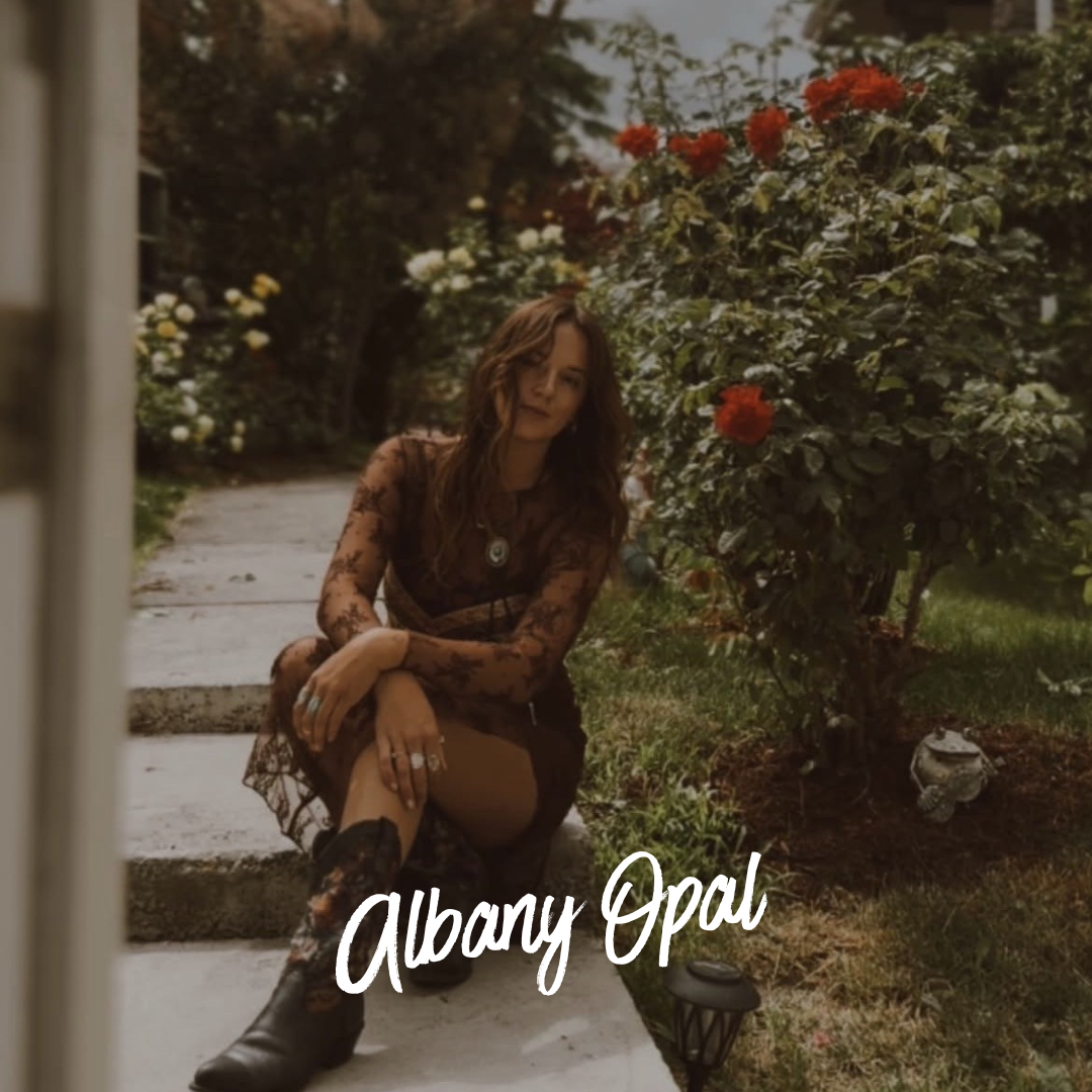 Woman in a black lace dress sits on a garden path surrounded by red roses; text reads “Albany Opal.”.
